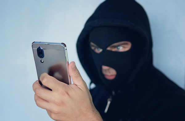 A scammer holding a cell phone while wearing a ski mask