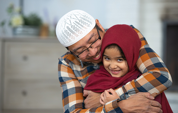 A father hugging and smiling with his young daughter who is wearing a Hijab.