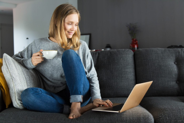 Image d’une femme heureuse confortablement installée dans un divan; elle sirote une boisson tout en consultant son ordinateur portable.