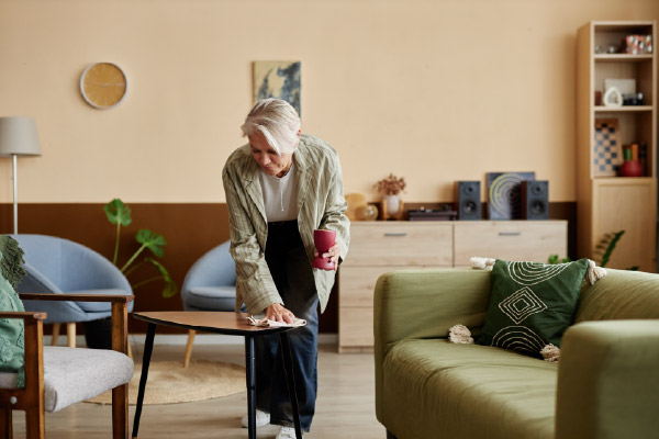 Person cleaning a coffee table in a modern apartment living room featuring a green sofa, armchairs, wood furniture, indoor plants, and contemporary decor.