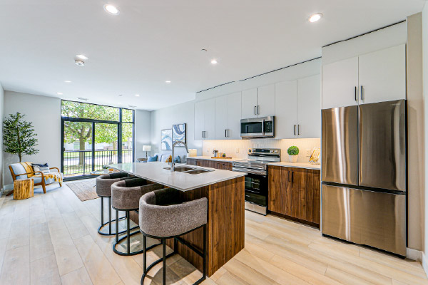 Modern open-concept apartment kitchen and living area featuring a large island with seating, white and wood-tone cabinetry, stainless steel appliances, floor-to-ceiling windows, and abundant natural light.