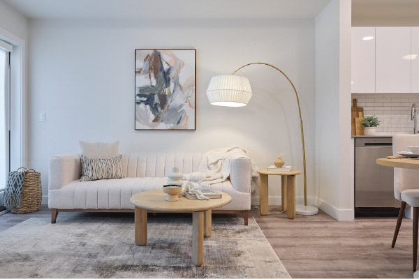 Modern apartment living room featuring a light-coloured sofa, round wood coffee table, floor lamp, abstract wall art, and an open-concept layout leading to a contemporary kitchen with white cabinetry. 