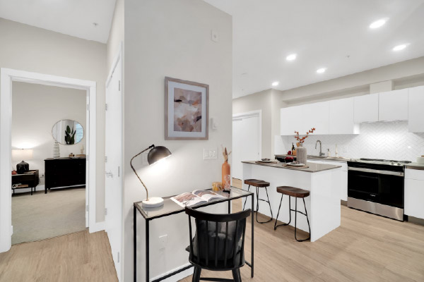  Open-concept apartment with a home office nook, modern kitchen featuring a large island and white cabinetry, and a doorway leading to a bedroom. 