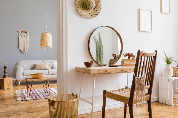 Bright apartment corner featuring a wooden vanity table with a round mirror and woven chair, complemented by neutral decor, indoor plants, and a cozy living area with a sofa, coffee table, and woven wall accents.