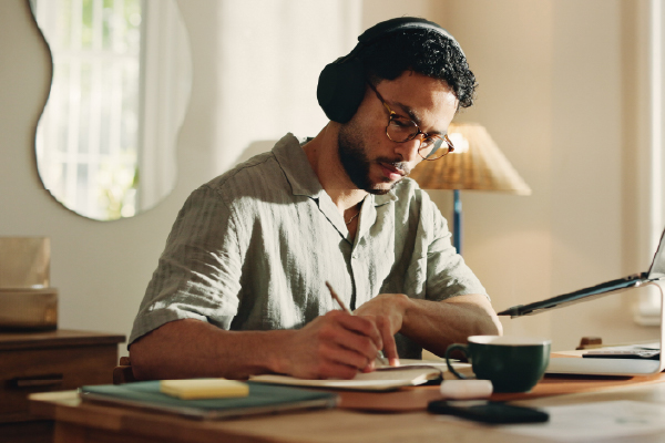 A man wearing headphones sits at a wooden desk in a bright home workspace, writing in a notebook beside a laptop on a stand. A coffee mug, sticky notes, and notebooks are arranged on the desk, with natural light streaming through nearby windows.