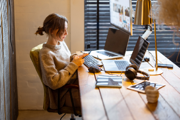 A young woman sits at a wooden desk, working on her phone while her laptop and desktop computer are open in front of her. On the desk are a plant, a lamp, an iPad, and headphones. She sits comfortably as she works. In the background, a wood plank wall and a window with closed blinds create a warm, enclosed atmosphere.